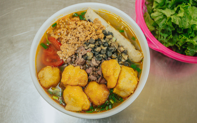 The first dish to mention is Hanoi snail noodle soup. The broth is prepared using snail stock and tomatoes. This dish includes vermicelli cooked in boiling water, accompanied by fresh vegetables, fish cakes, fried tofu, finely chopped scallions, and snails. It can be enjoyed with a squeeze of lime, shrimp paste, or chili sauce. Photo courtesy of Xuan Phuong